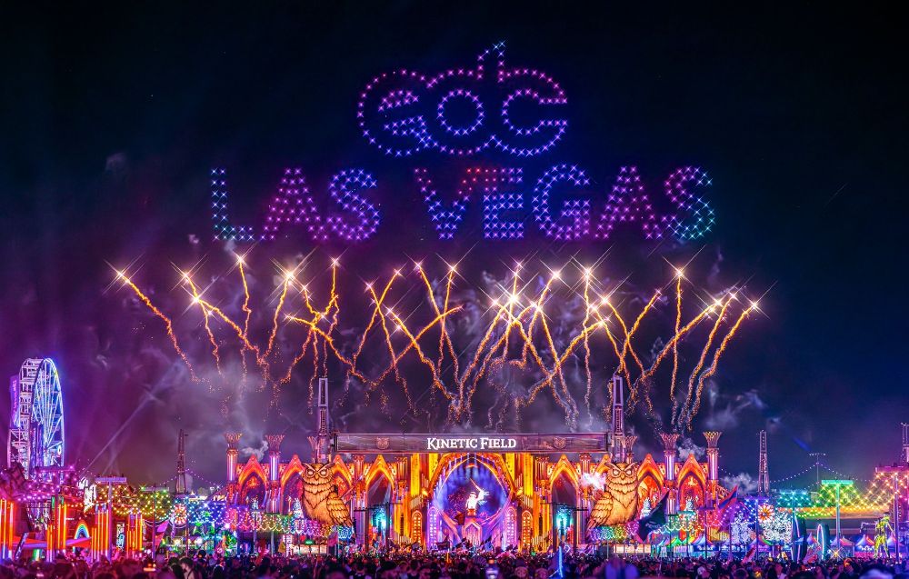 : "A vibrant nighttime scene at EDC Las Vegas featuring a dazzling display of fireworks above the Kinetic Field stage. The words 'EDC Las Vegas' are illuminated in the sky with glowing, colorful lights. The stage is adorned with whimsical, festival-themed decorations, including neon lights, owl sculptures, and lively structures, while a large Ferris wheel glows in the background. A crowd of festival-goers enjoys the energetic atmosphere under the night sky, surrounded by a kaleidoscope of colors and electric vibes."