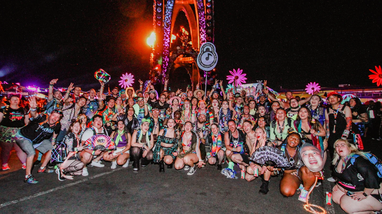A massive group of festival-goers dressed in vibrant rave outfits posing together at night, surrounded by neon lights, glowing decorations, and the electrifying energy of the event