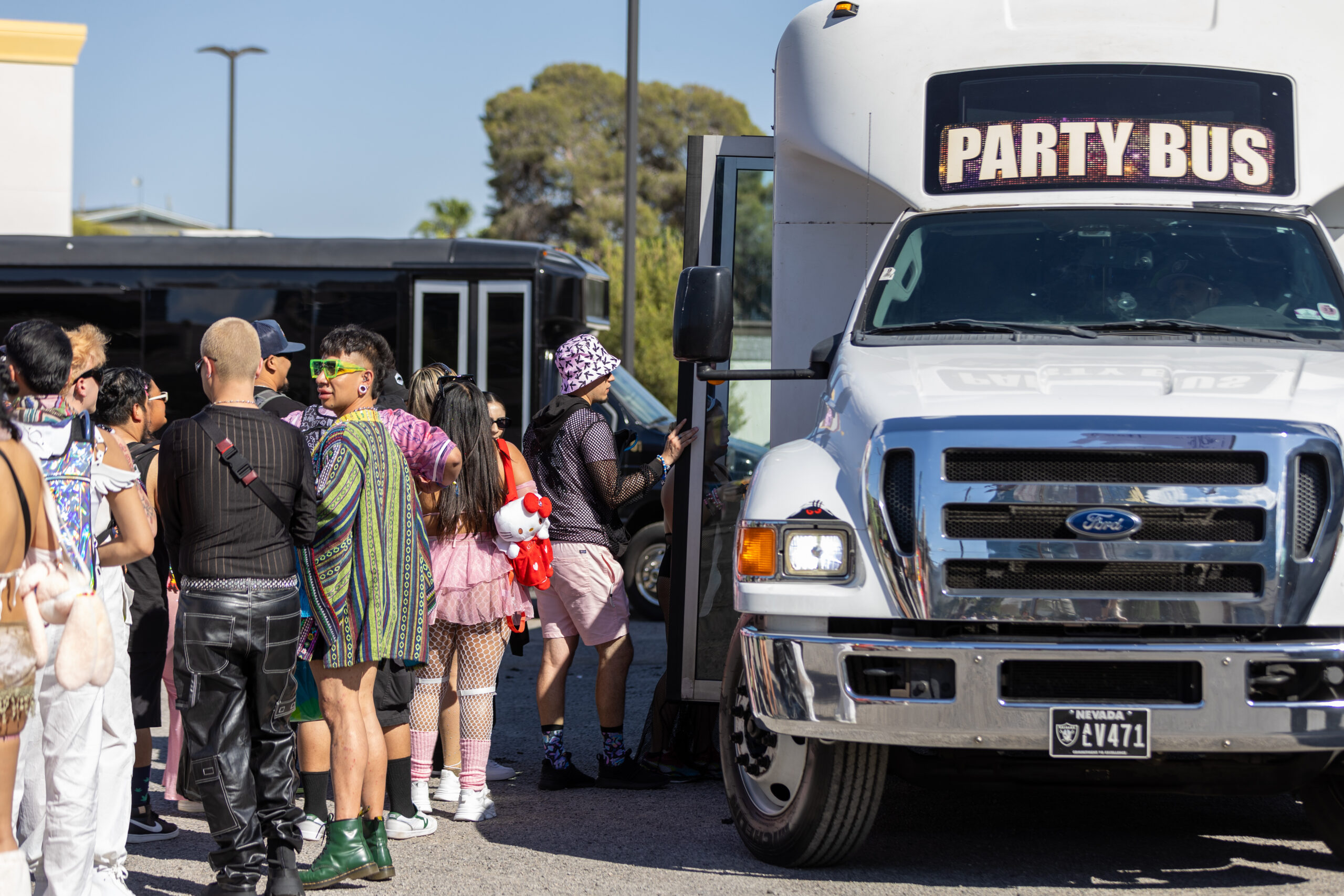 A group of colorfully dressed festival-goers lines up to board a Party Bus, with standout outfits, accessories, and a Hello Kitty plush backpack visible.